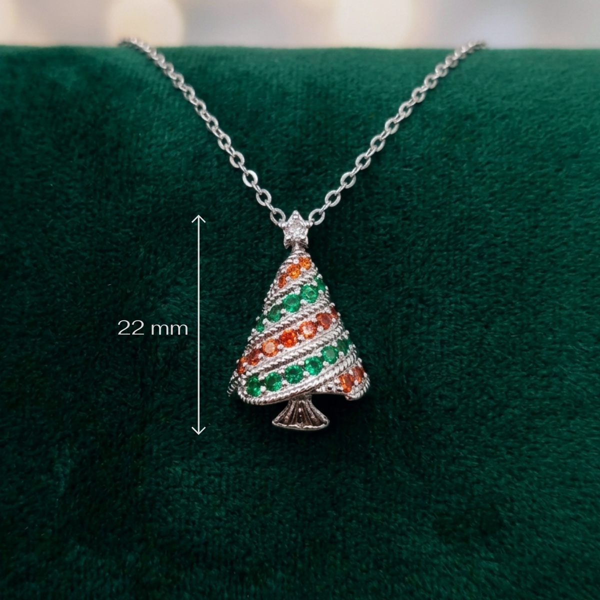 A silver Christmas tree pendant necklace with green and orange stones, displayed on a dark green velvet surface with a '22 mm' size measurement overlay.
