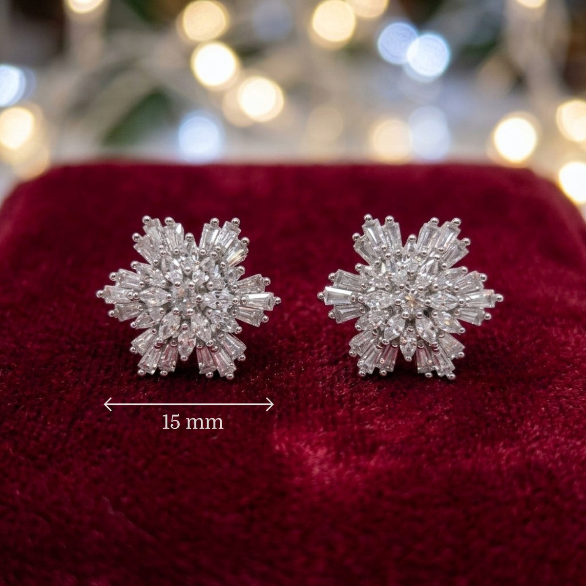 A pair of snowflake-shaped stud earrings made of clear baguette-cut gemstones in a silver-toned setting, displayed on a deep red velvet surface with a blurred festive bokeh background. A dimension marker indicates 15 mm.