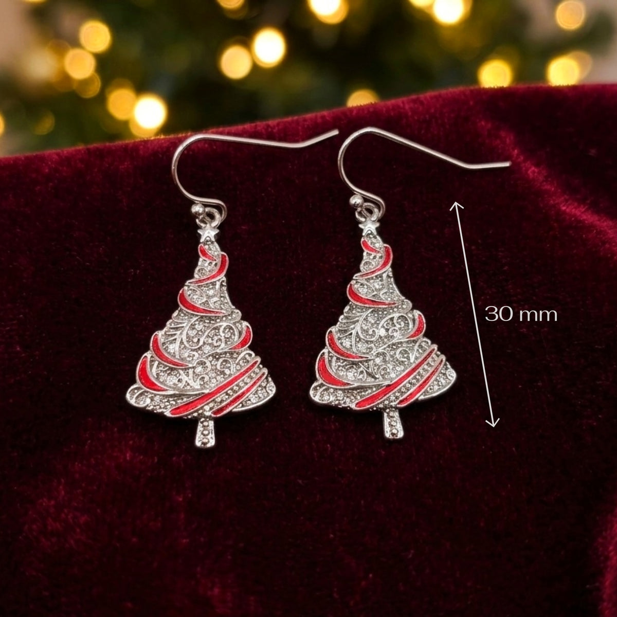 A pair of silver-toned dangle earrings shaped like Christmas trees with red enamel accents, displayed on a dark red velvet surface with a blurred festive background and a '30 mm' size marker.