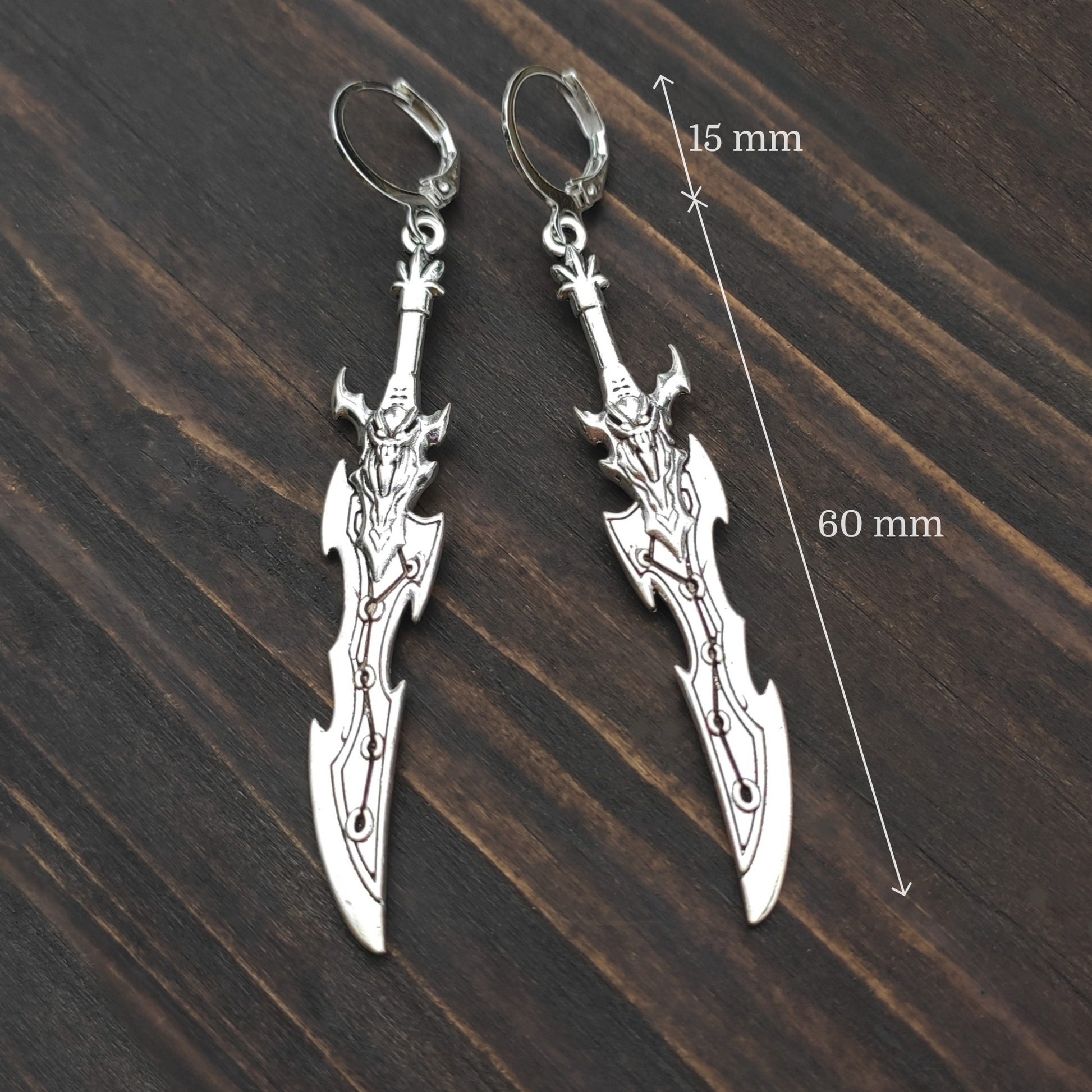 Silver sword earrings on a wooden surface with measurements indicated.
