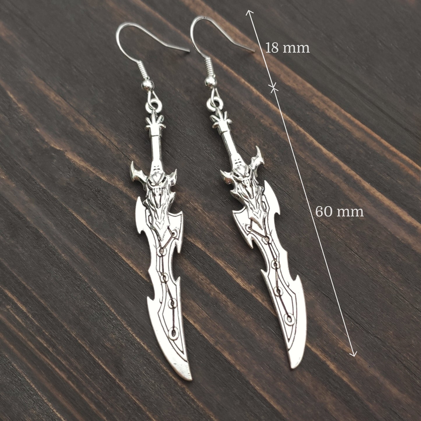 Silver sword earrings on a wooden surface with measurements indicated.