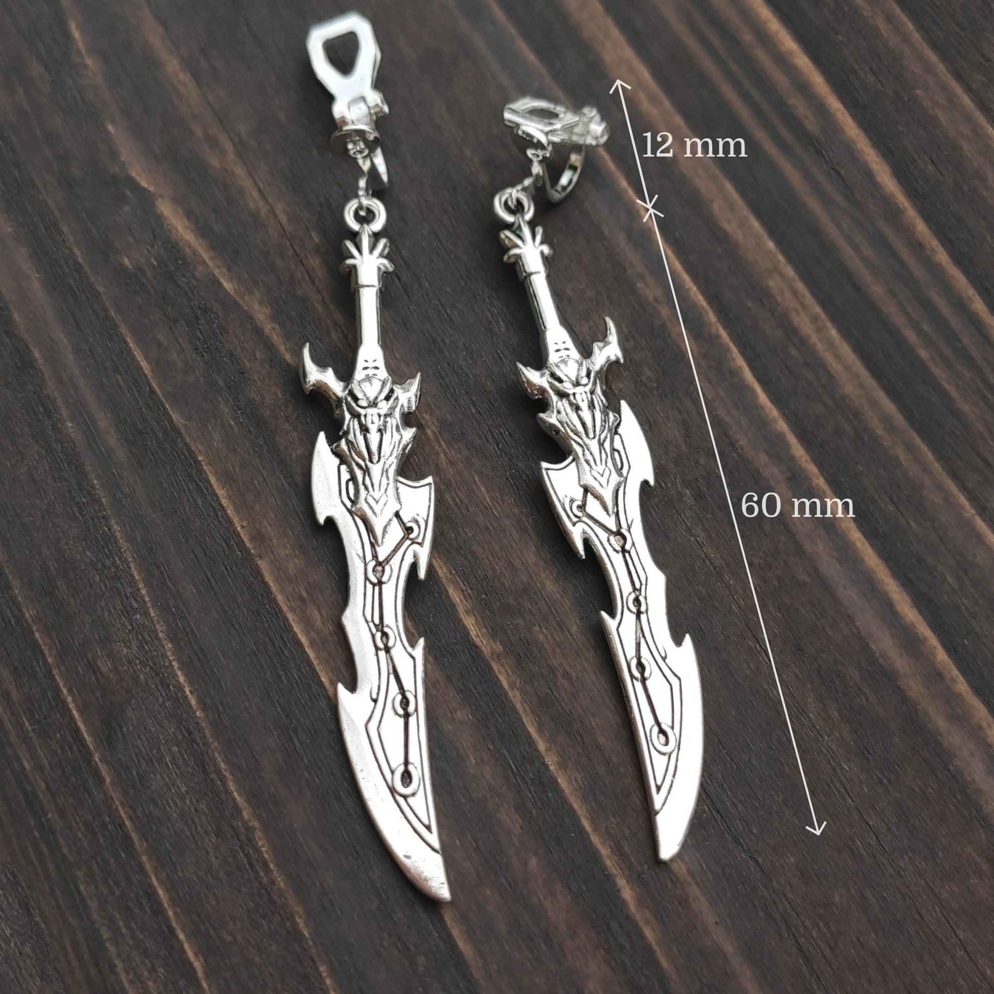 Silver sword earrings on a wooden surface with measurements indicated. The earrings feature clip-ons for unpierced ears.