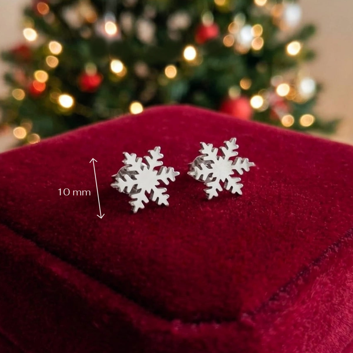 Dainty snowflake stud earrings made from stainless steel on a red velvet background with festive lights in the background. A measurement scale reads 10mm