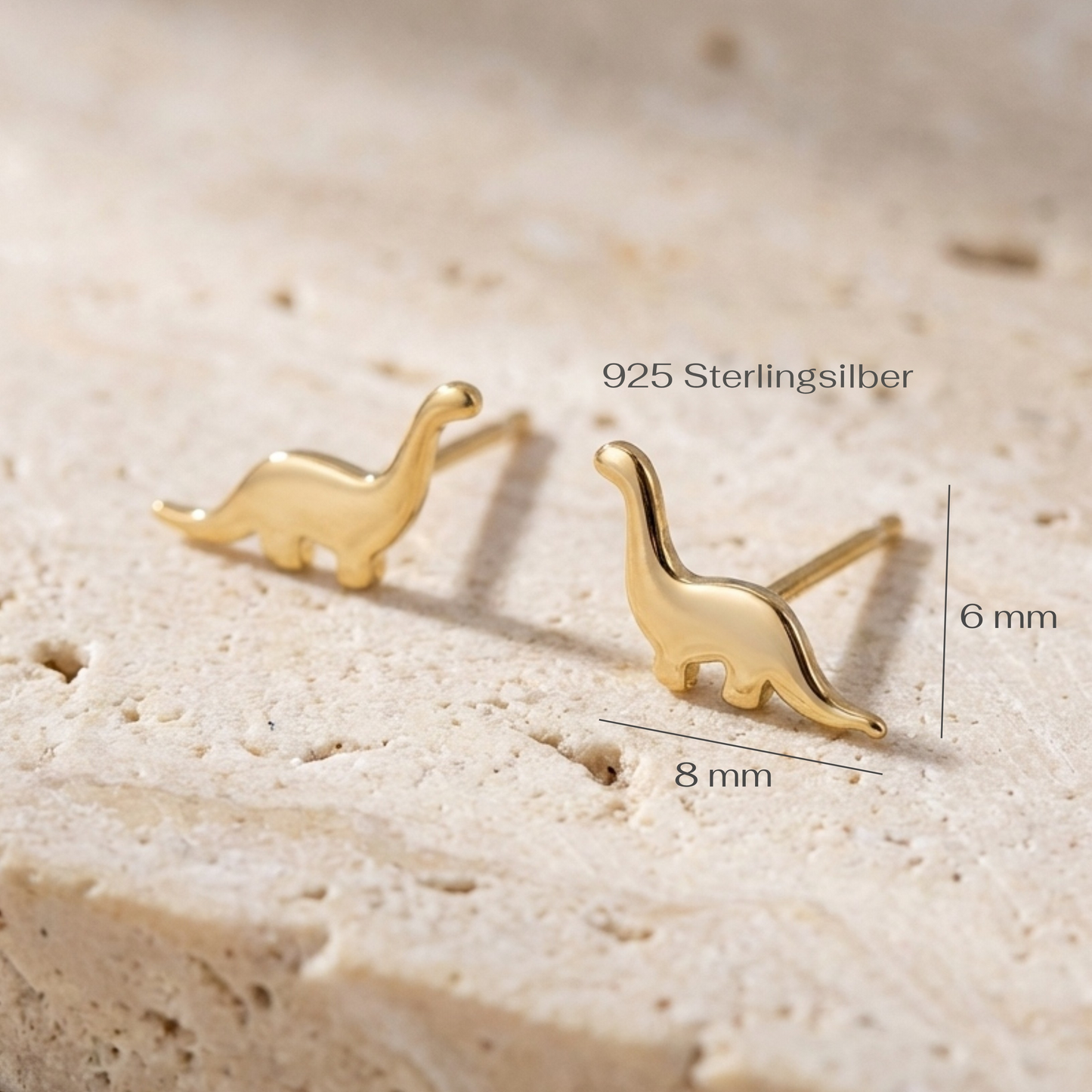 Gold dinosaur-shaped earrings on a beige stone surface with measurements.