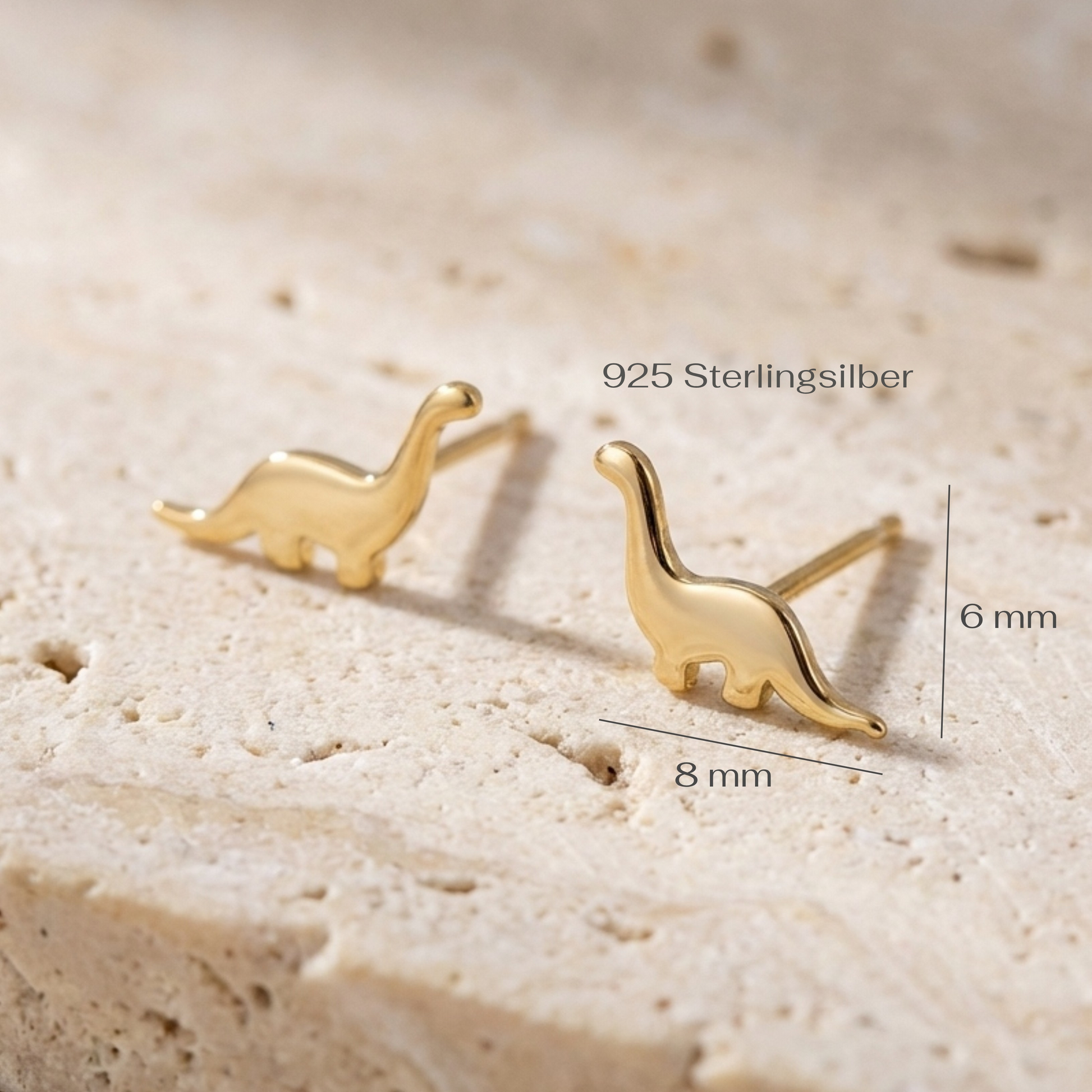 Gold dinosaur-shaped earrings on a beige stone surface with measurements.