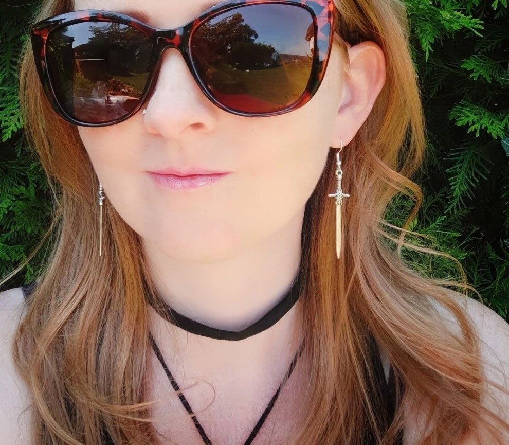Close-up of a person wearing sunglasses and silver sword earrings with a blurred outdoor background